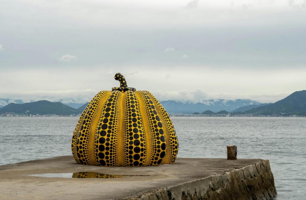 Yellow pumpkin sculpture by Yayoi Kusama on Naoshima Island pier, with sea and mountains in background.