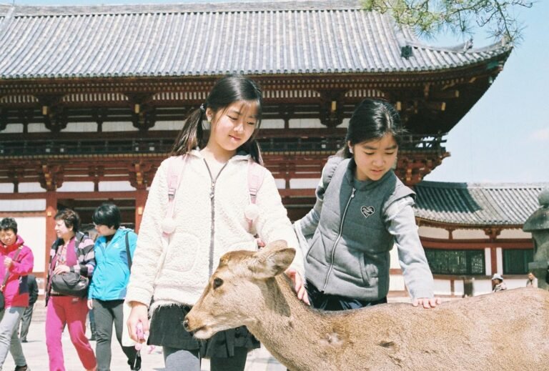 Two young girls petting a deer outside a traditional Japanese temple in Nara.